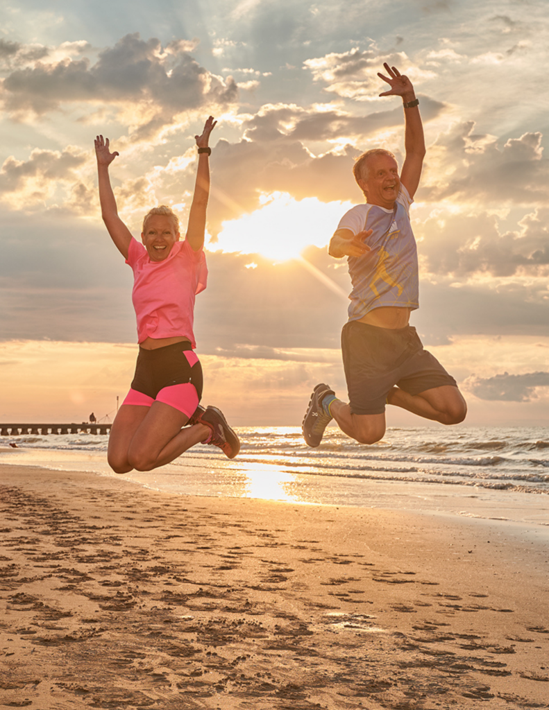 Béatrice Drach-Schauer und Johannes Drach springen jubelnd in die Luft am Strand bei Sonnenuntergang – Symbolbild für Energie, Fitness und Leistungsfähigkeit ab 45